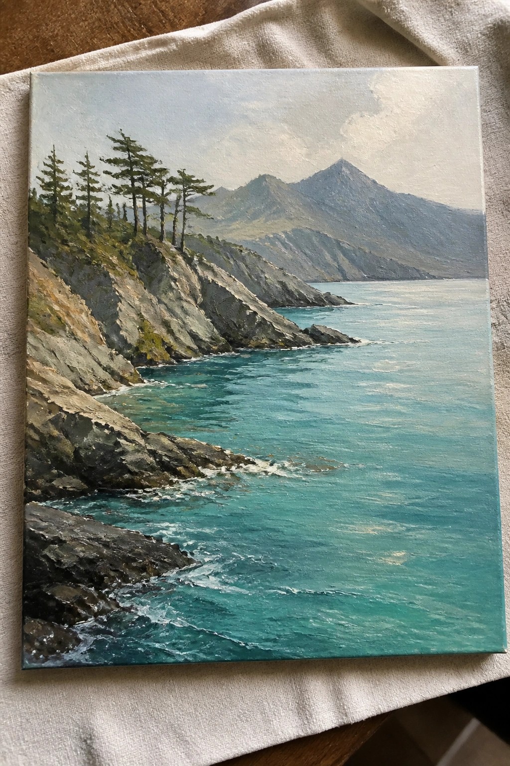 Realistic oil painting of rocky cliffs with pines above turquoise waves and misty mountains on white cloth.