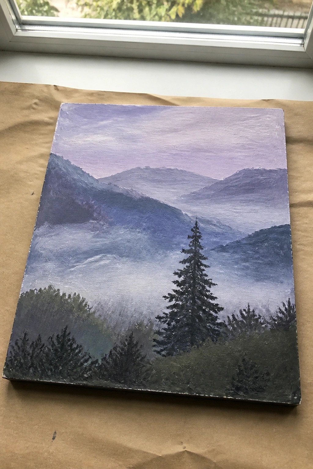 Misty purple and blue mountain landscape painting with pine trees on canvas under window.