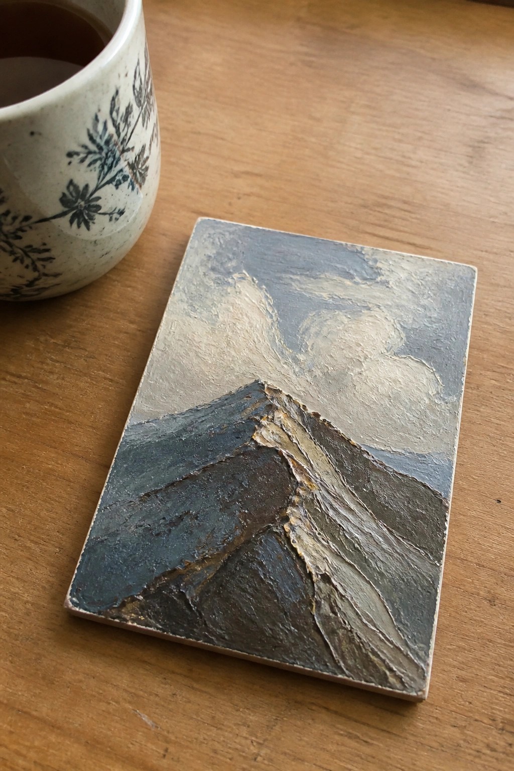 A small rectangular oil painting of a textured mountain range in dark and light tones rests on a wooden table next to a ceramic mug with a floral pattern.
