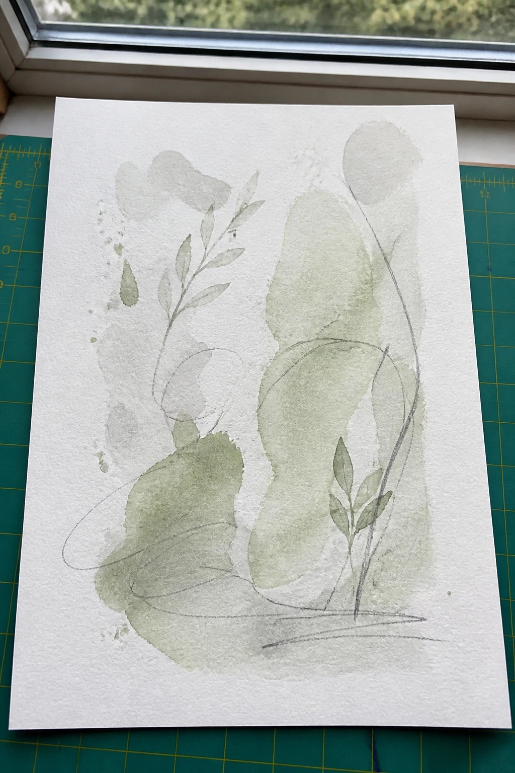 Abstract green watercolor leaves, stems, and organic shapes on white paper by window