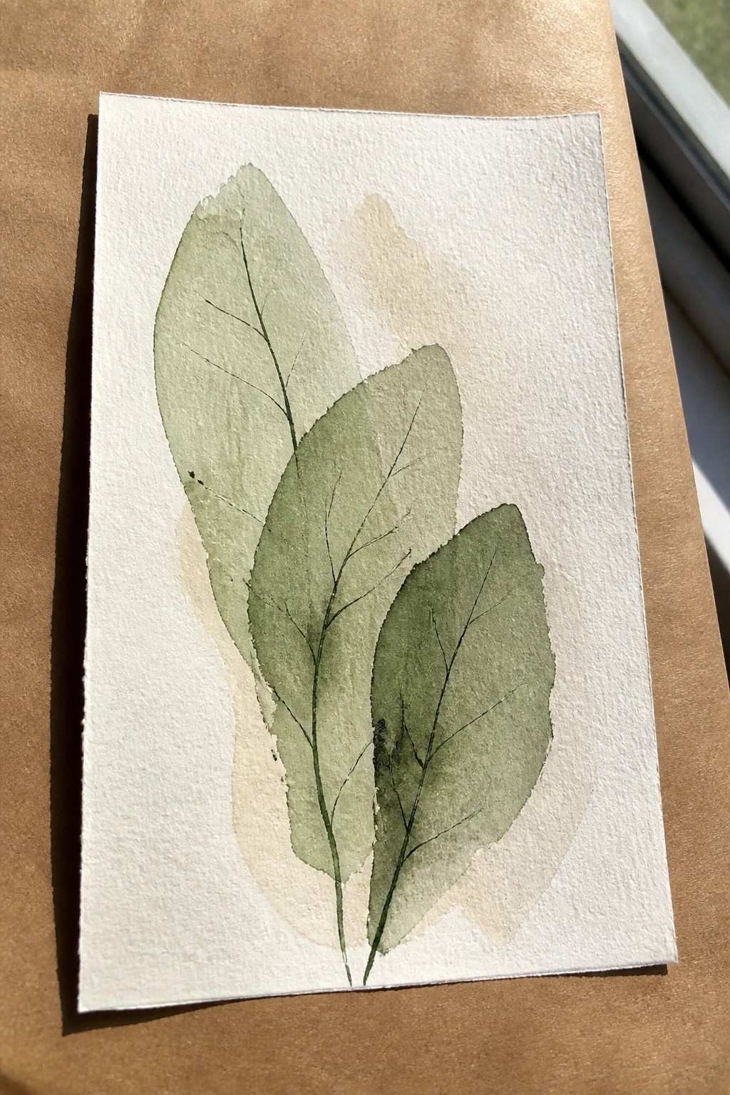 Three overlapping green watercolor leaves with veins on textured white paper over brown