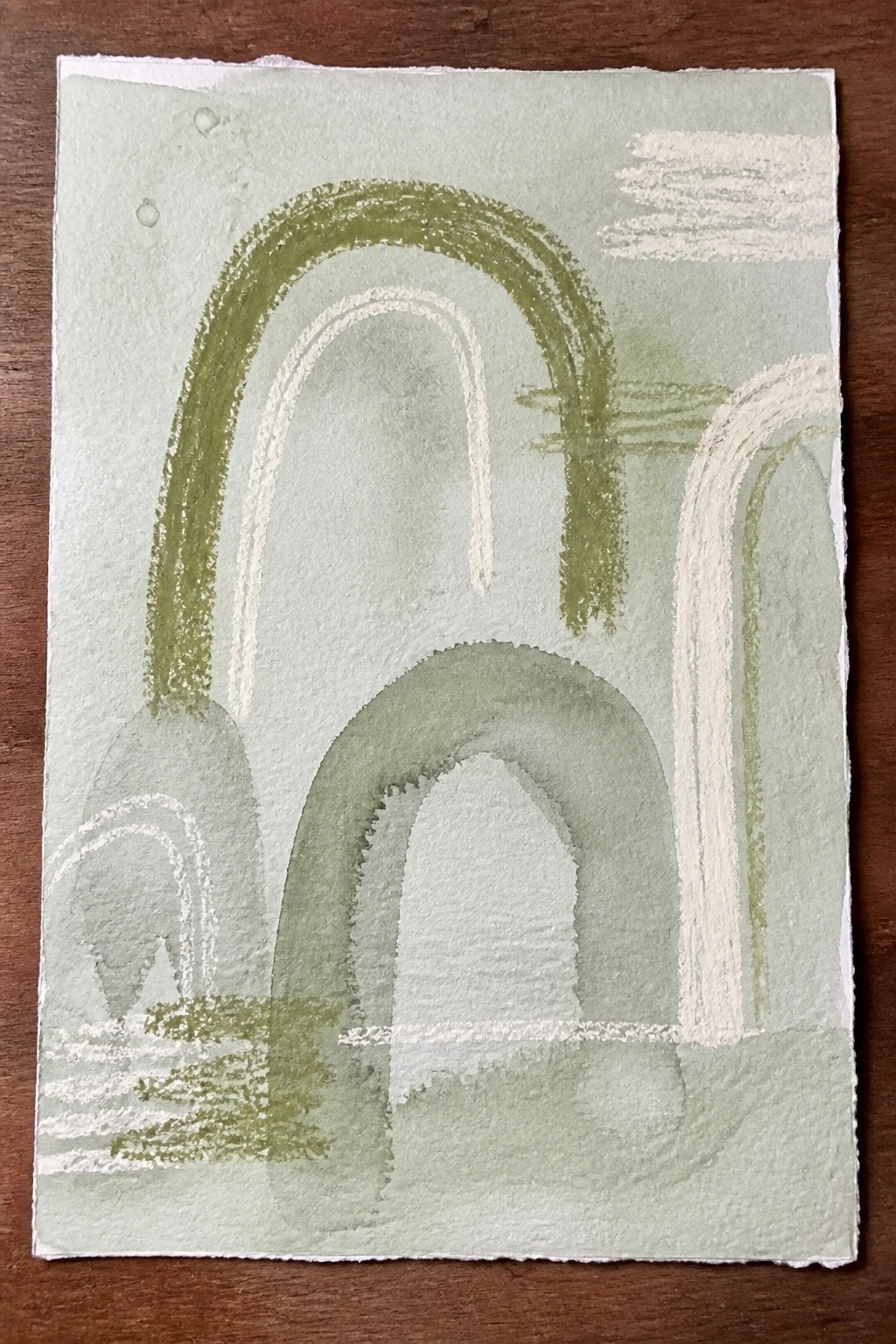 An abstract painting featuring multiple green arches and curved shapes on a light green background with white brushstrokes, placed on a wooden surface.