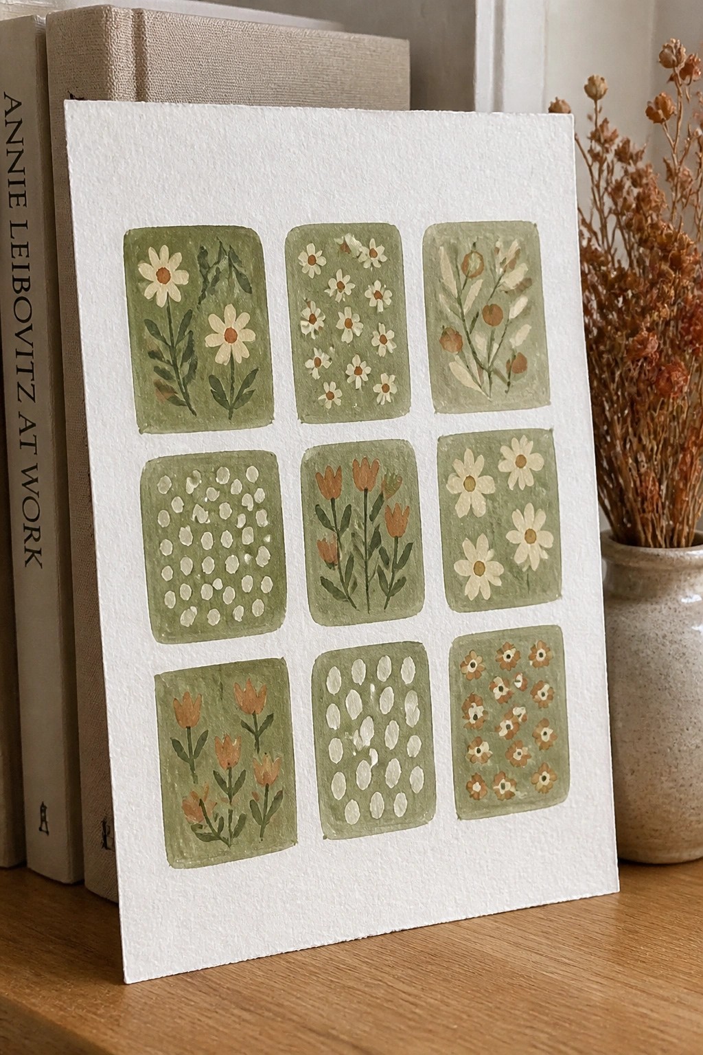 Watercolor artwork with nine green floral panels on white paper beside books and dried flowers