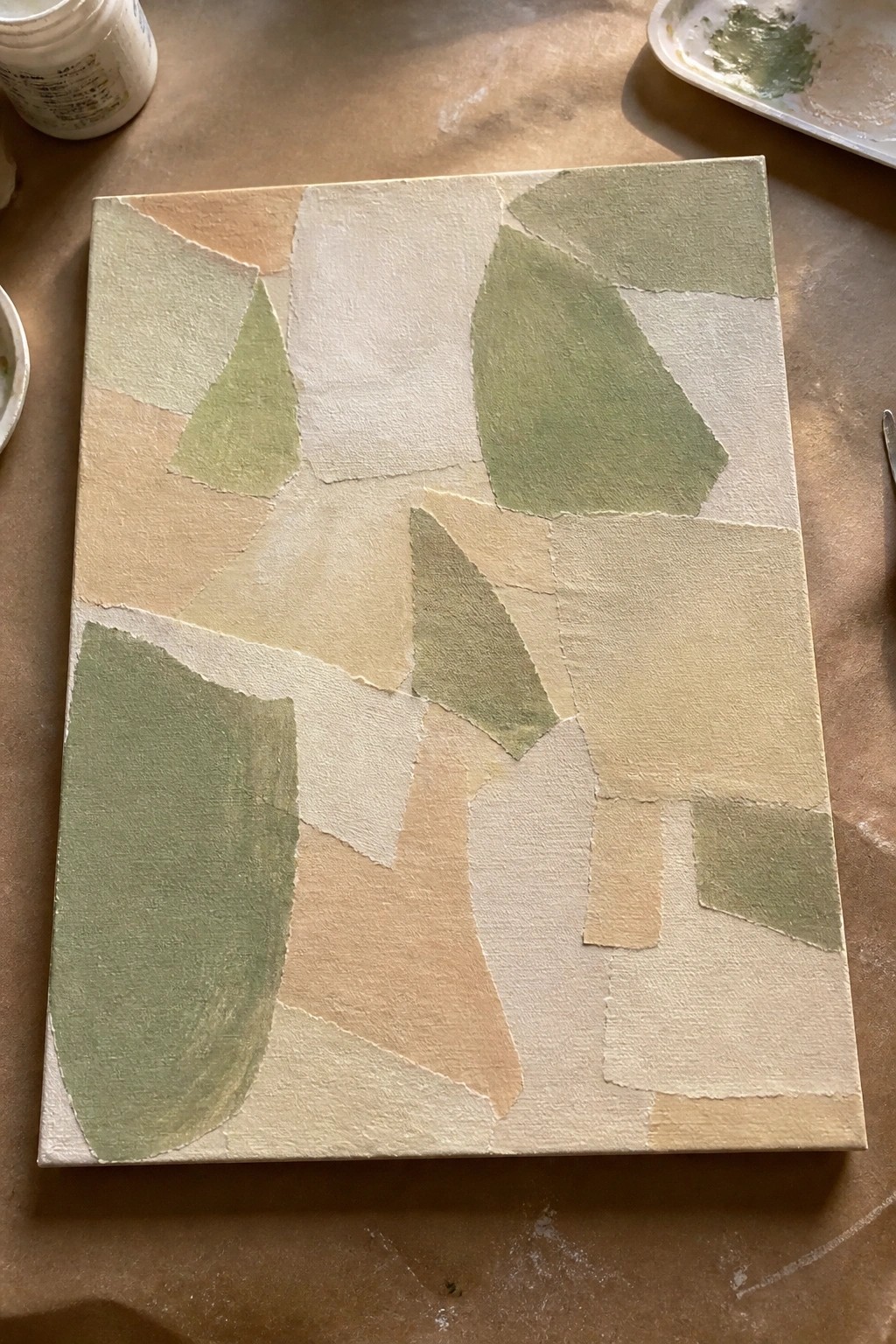 Abstract canvas with irregular green and beige shapes amid paint jars and palette on wood.