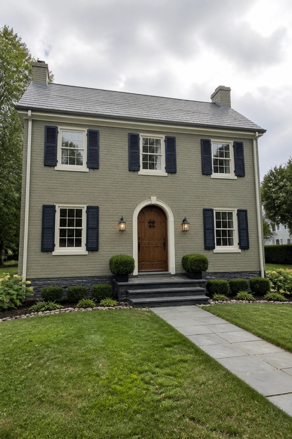 Two-story gray brick house with dark blue shutters, arched wooden door, and stone steps.
