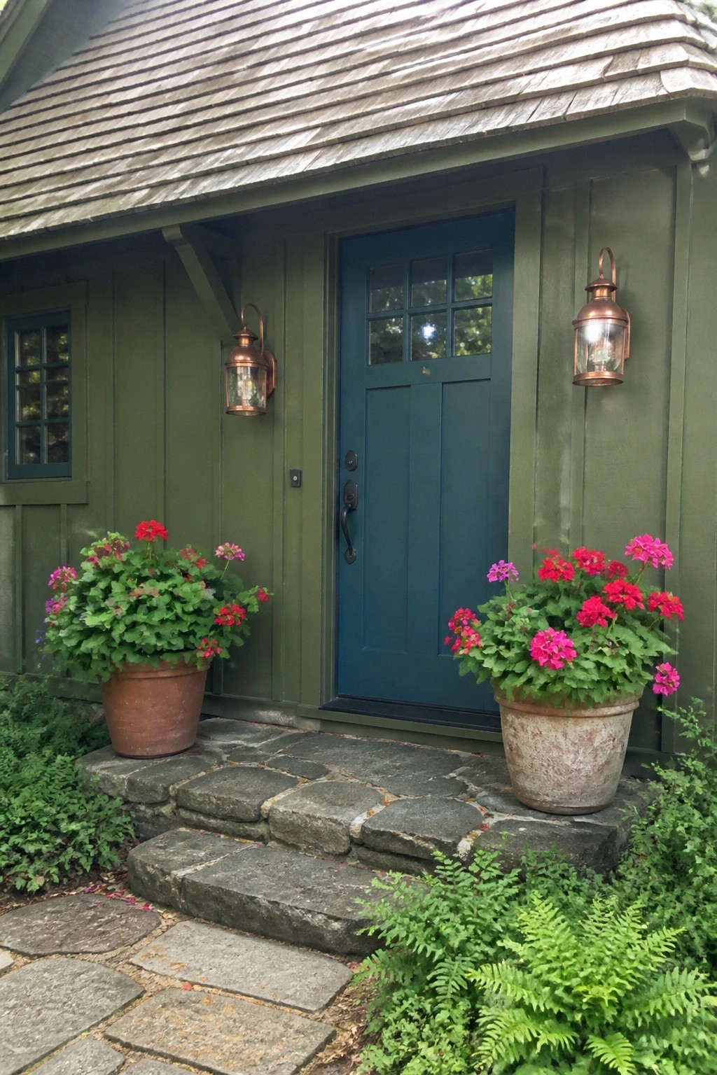 Green house with teal door, copper lanterns, and blooming flower pots on stone steps.