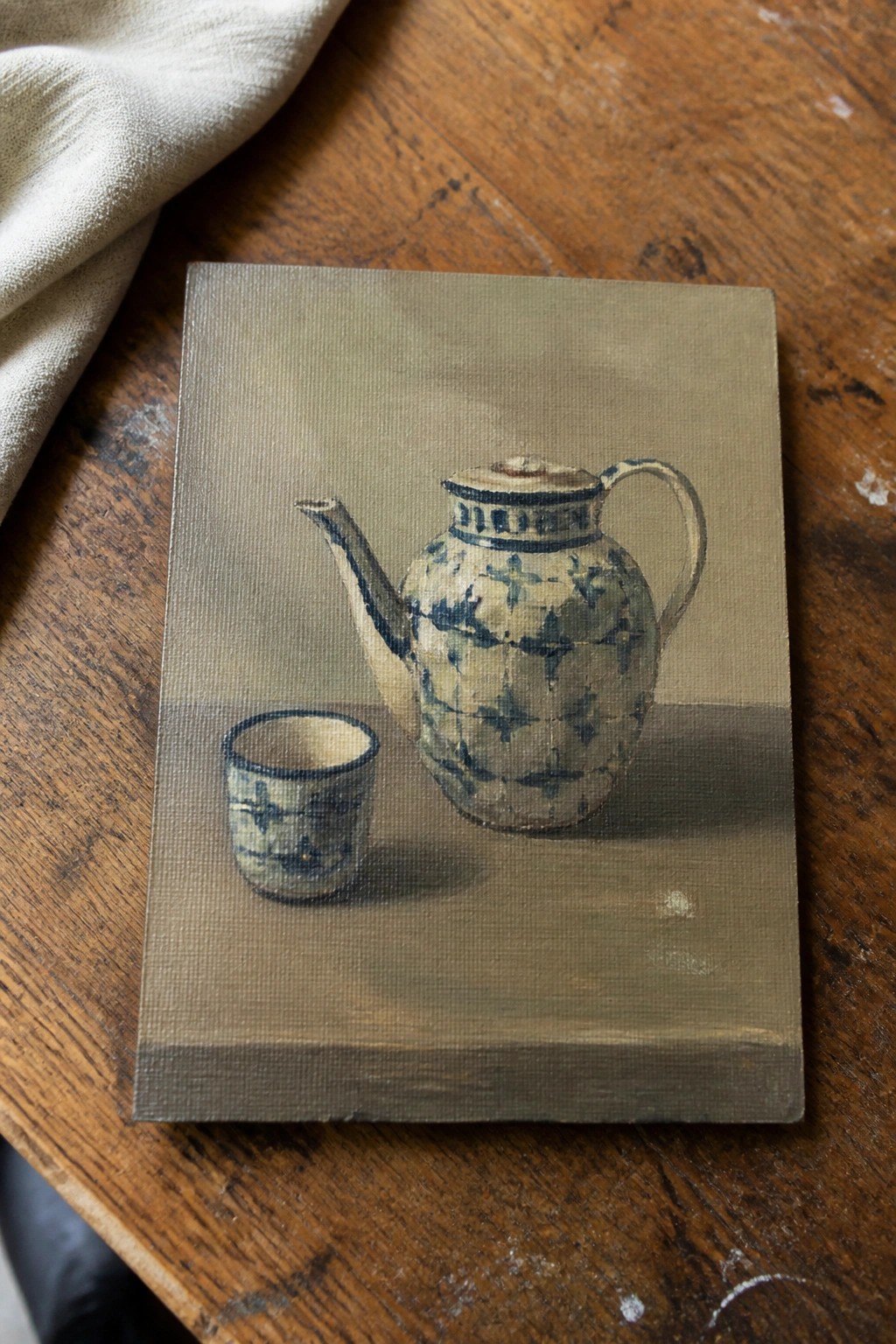 Realistic painting of blue-patterned teapot and cup on wooden table beside white cloth