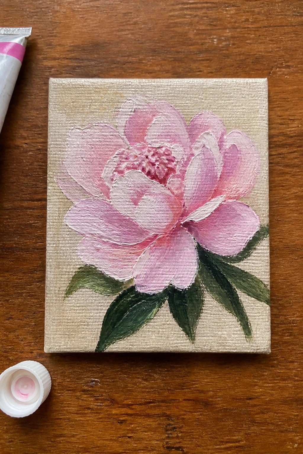 Small canvas with pink peony painting, paint tube, and cap on wood