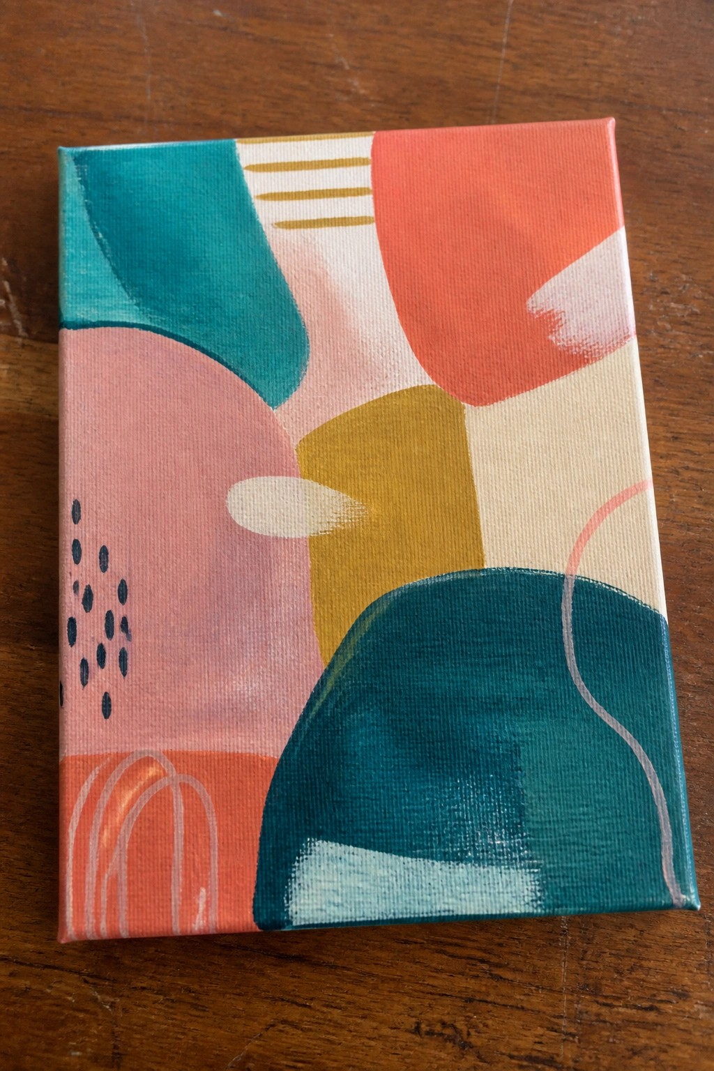 Abstract painting with teal, coral, pink, yellow shapes and dots on wood table