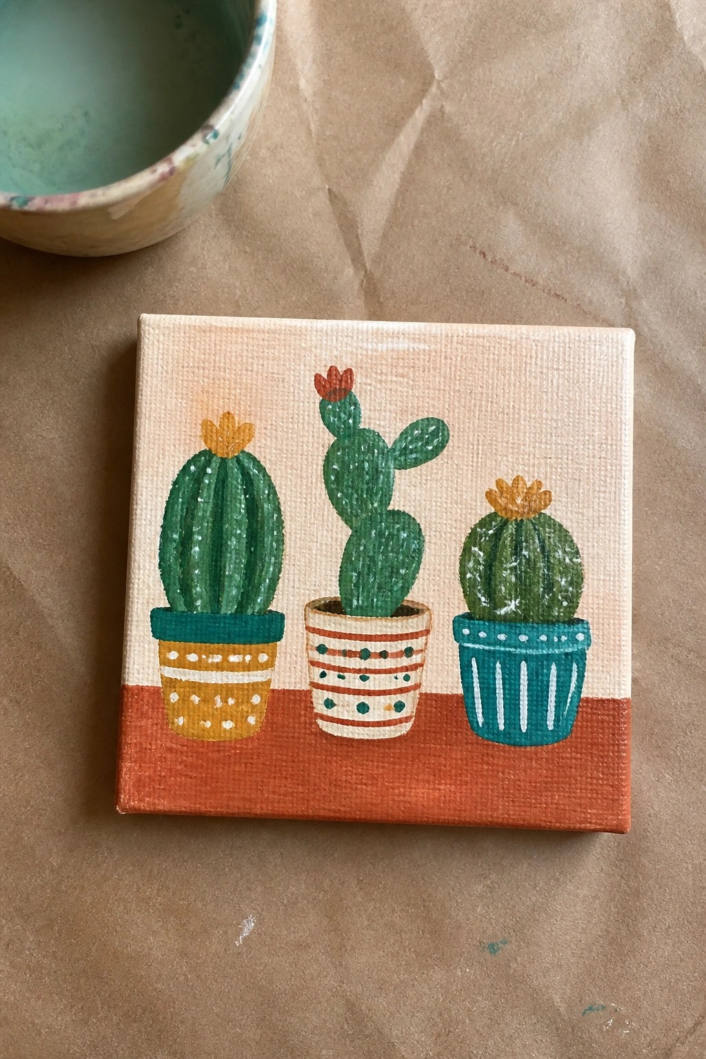 Three potted cacti painted on small canvas beside speckled bowl