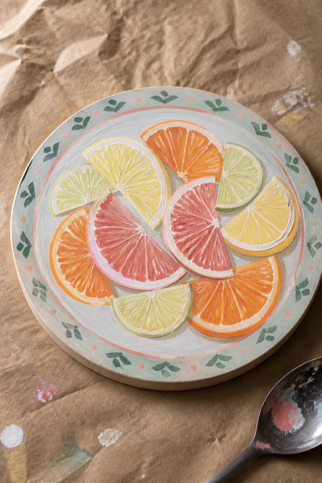 Painted wooden plate with sliced citrus fruits and leaf-patterned border beside spoon.