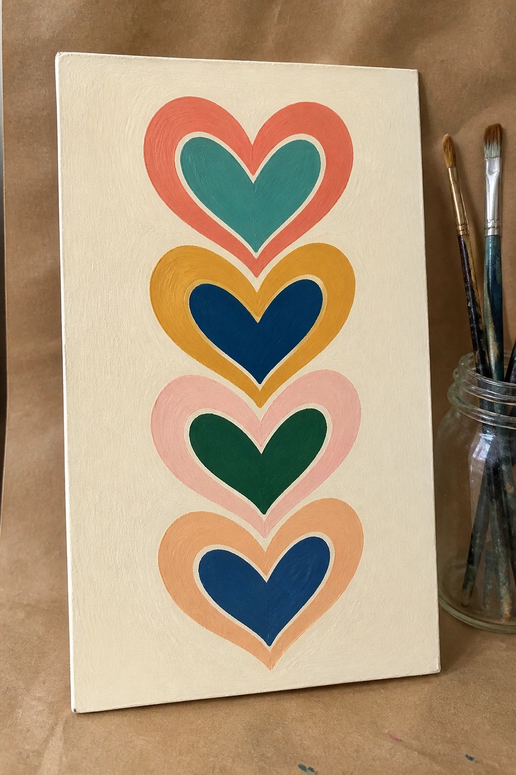 Four stacked colorful hearts painted on white canvas beside jar of brushes.