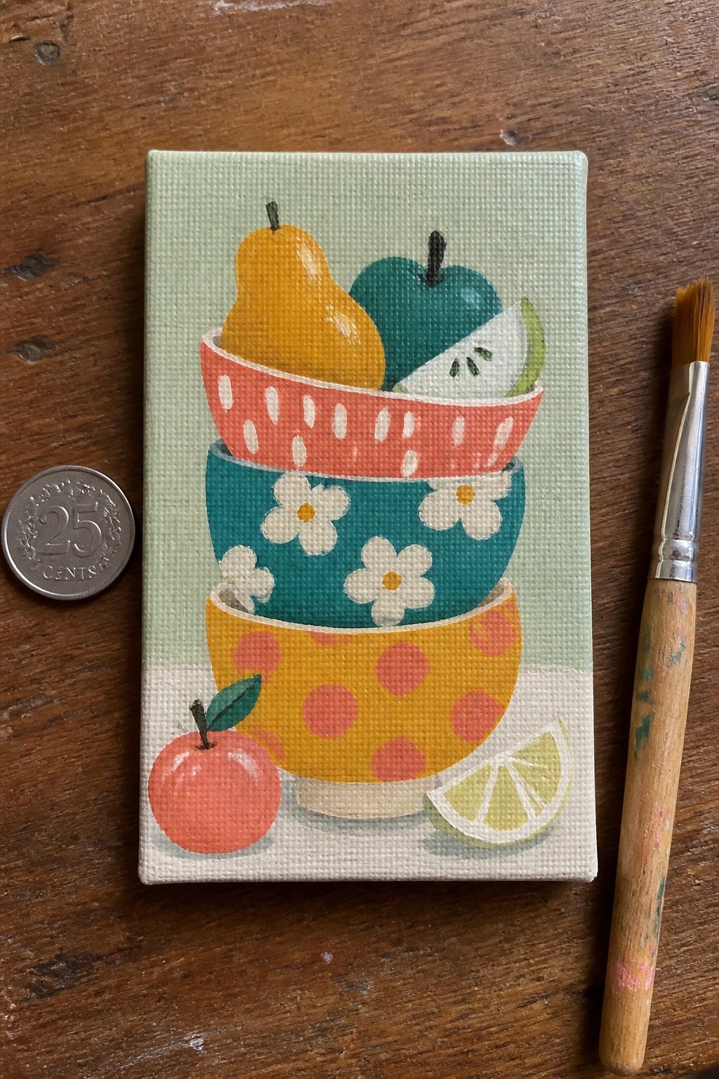 Small canvas painting of stacked colorful bowls and fruits beside quarter and brush