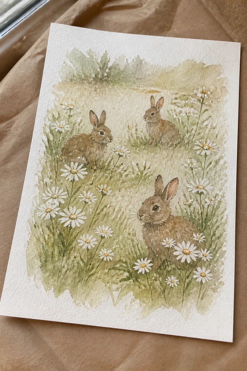 Watercolor painting of three brown rabbits among white daisies in a green meadow
