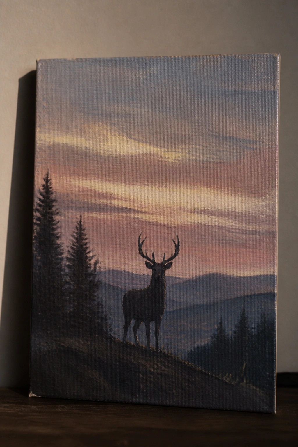 Silhouetted stag with antlers on a hillside beneath a colorful sunset sky with mountains and pines.