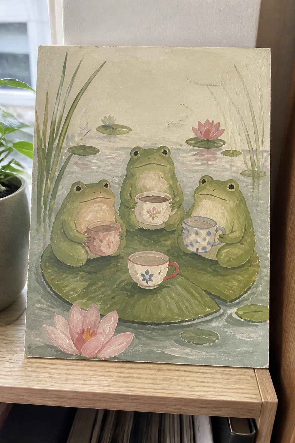 A painting of three green frogs sitting on lily pads in a pond, each holding a patterned teacup with water lilies and reeds around them.