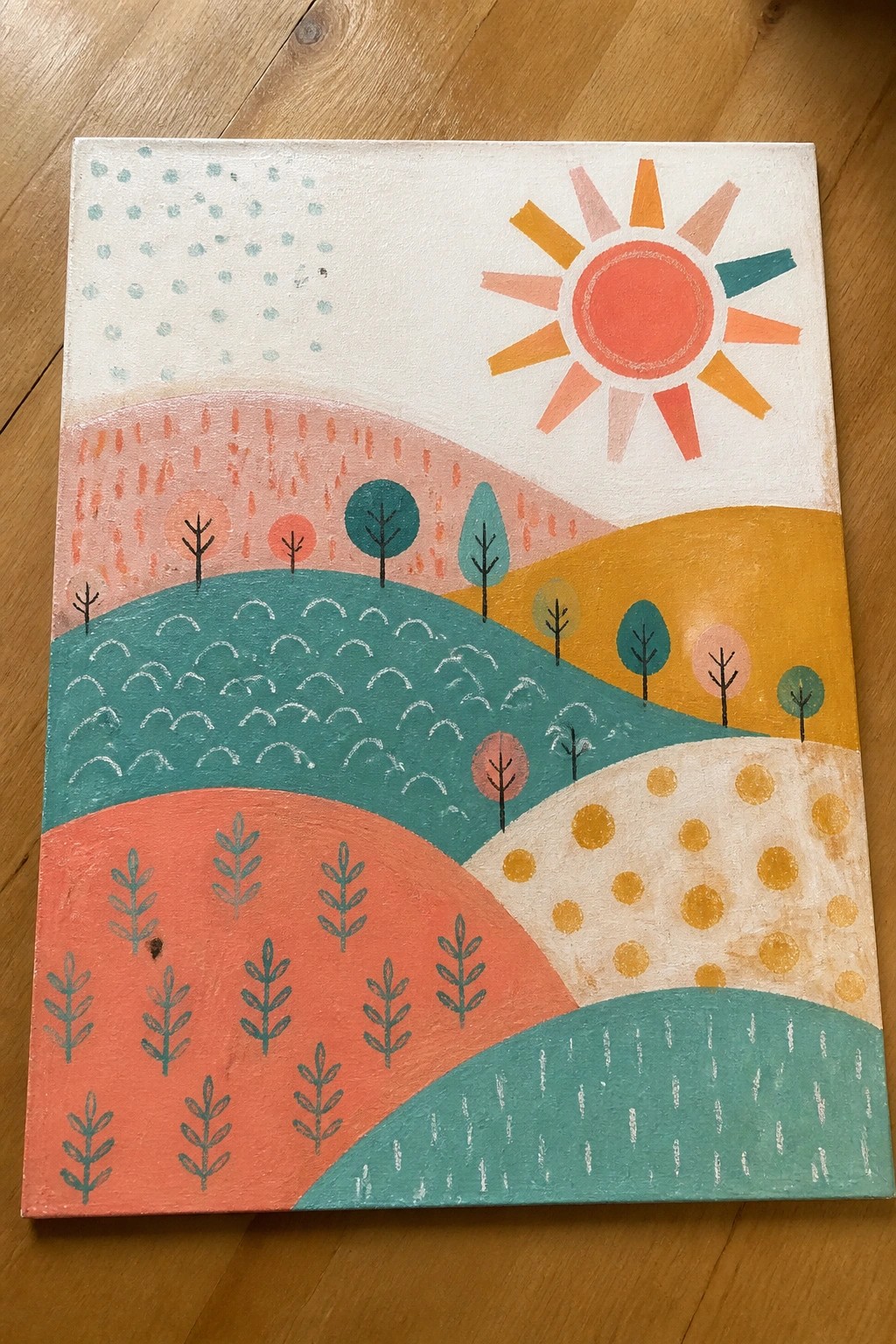 A painting of rolling hills in teal, orange, yellow, and pink with a red sun and stylized trees on a wooden table.