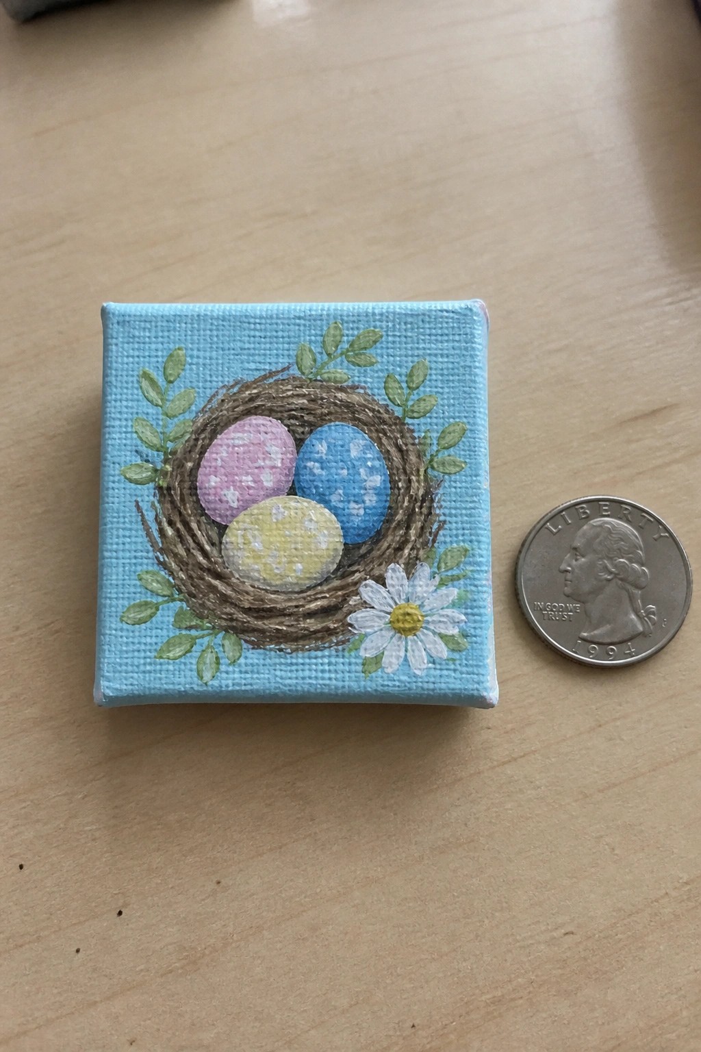 Small canvas painting of nest with pink blue yellow eggs beside quarter coin