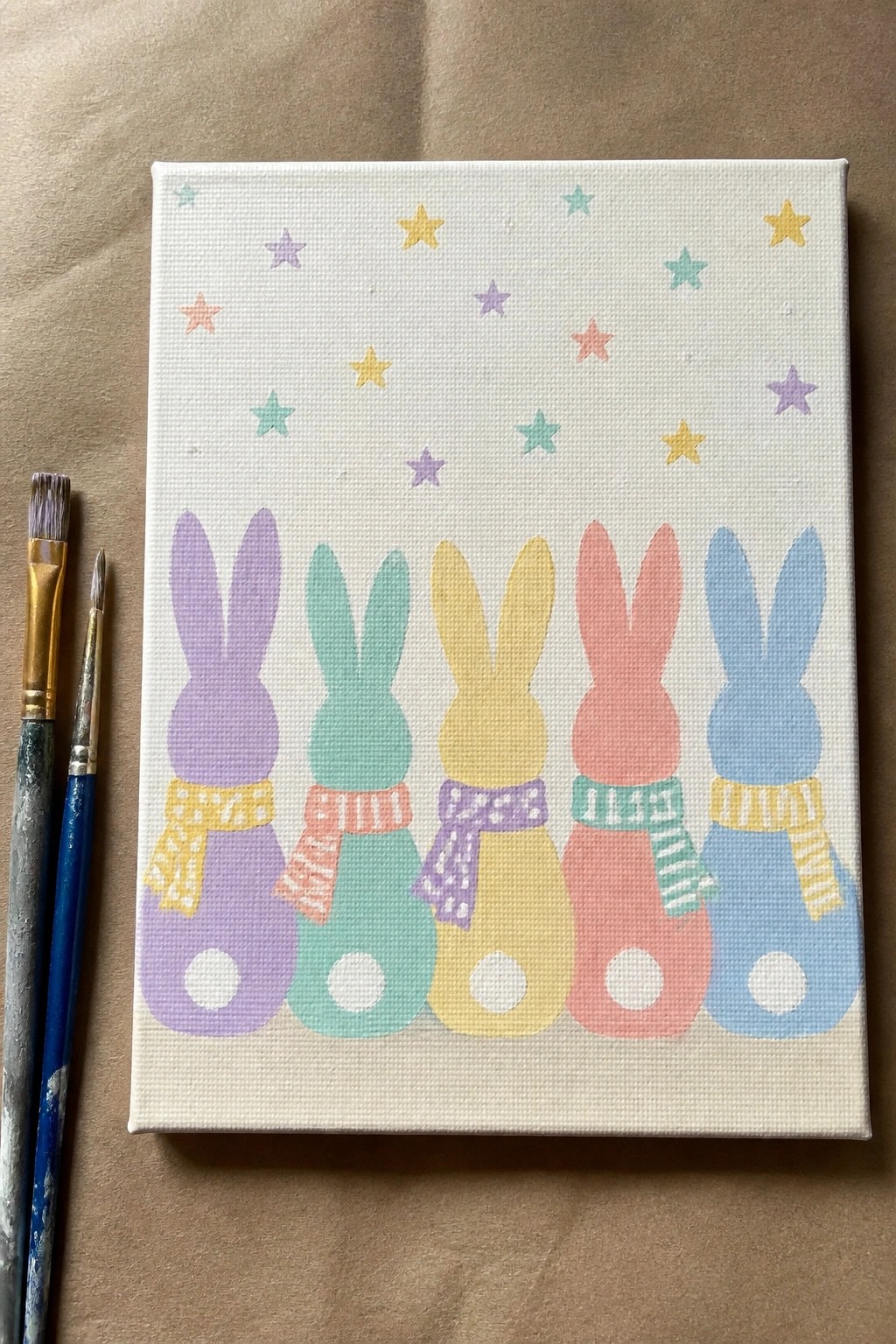 Five pastel bunnies in scarves with stars on canvas beside paintbrushes