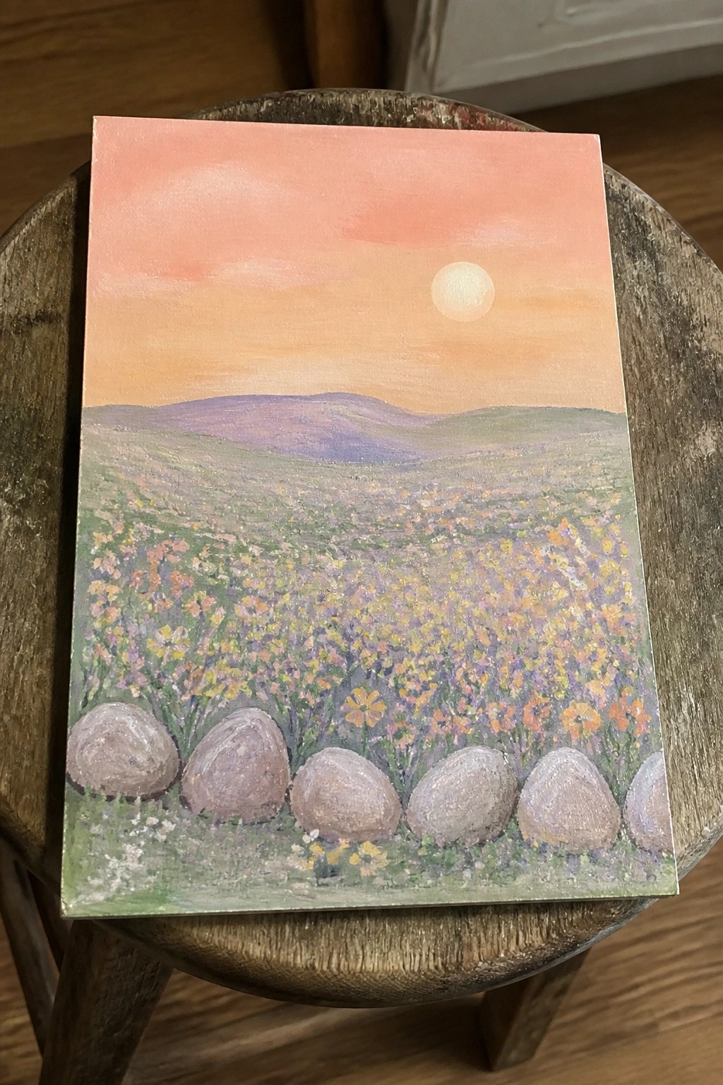 Painting of wildflower meadow with gray rocks under pink sunset sky on wooden stool