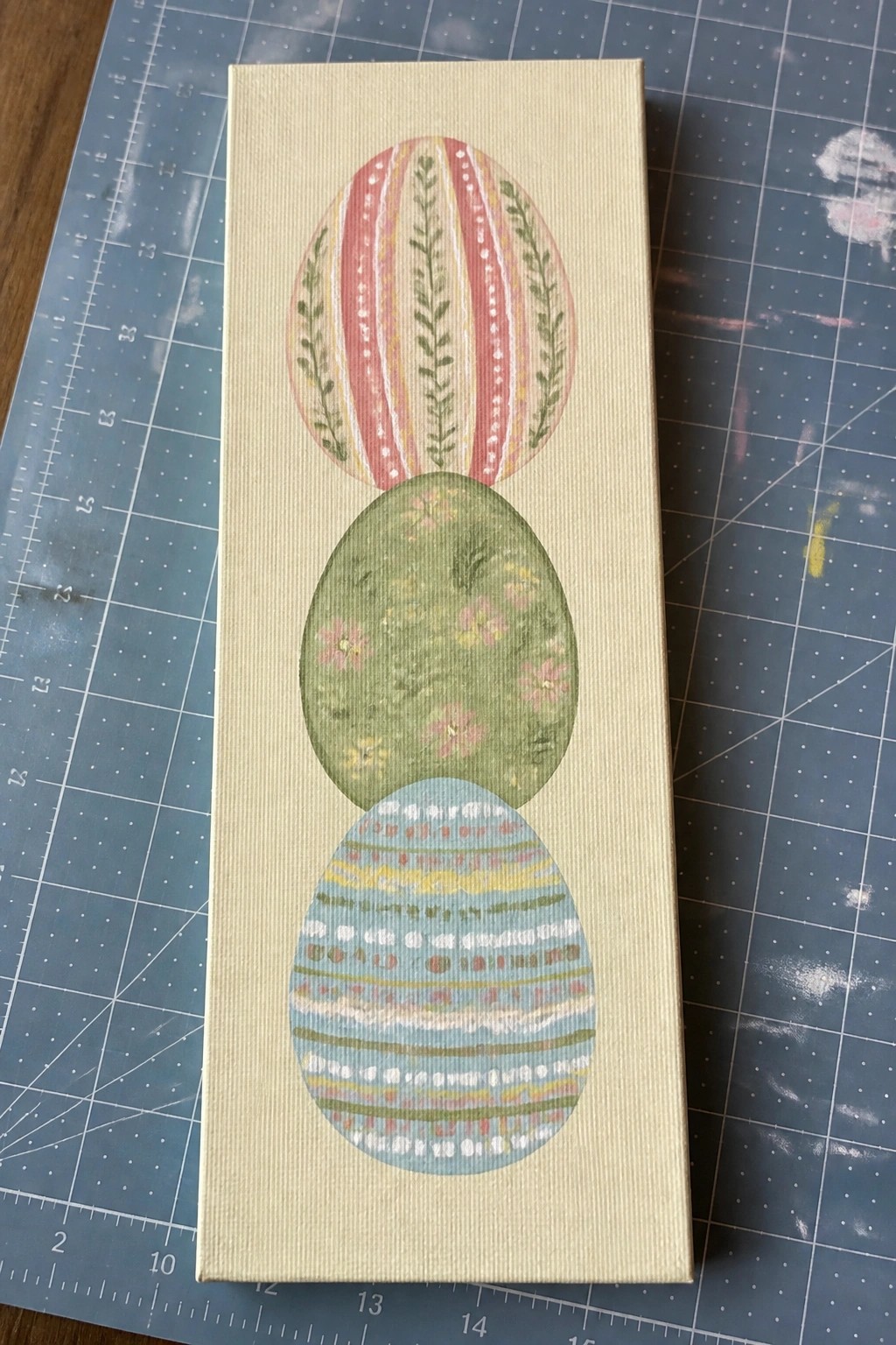 Three stacked pastel Easter eggs with floral and striped designs on canvas over blue mat.