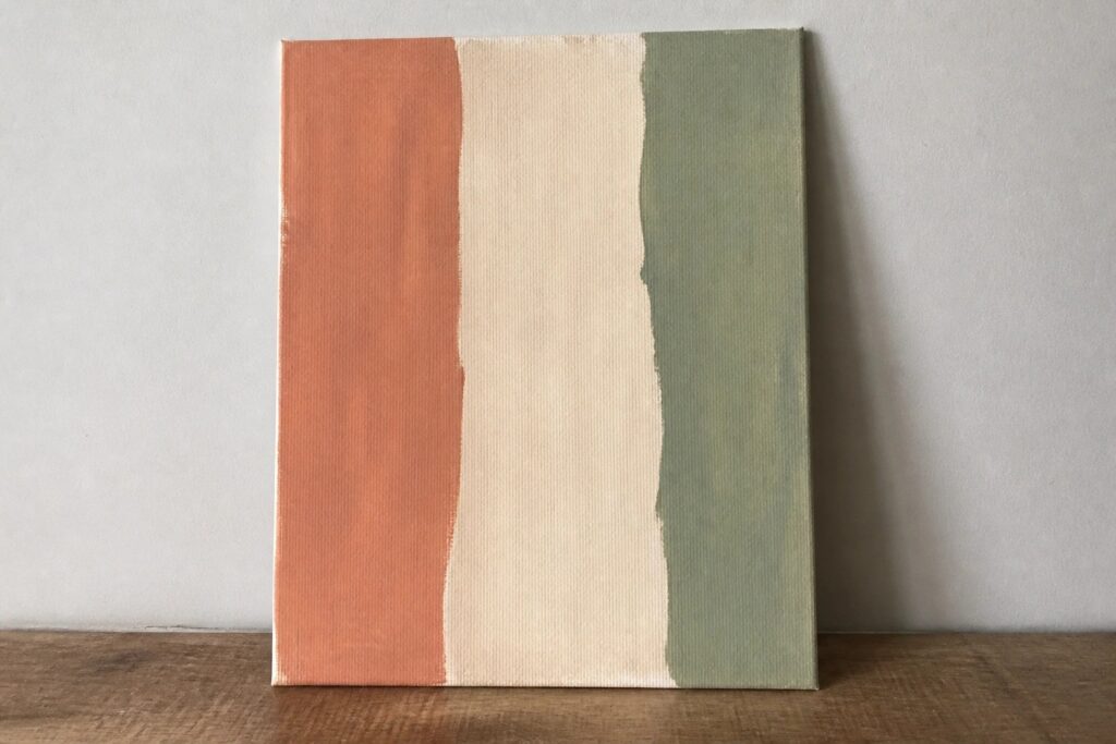 24 Easy Abstract Painting Ideas for Beginners Using Simple Color Blocks Abstract canvas with vertical orange, beige, and green stripes on wooden surface.