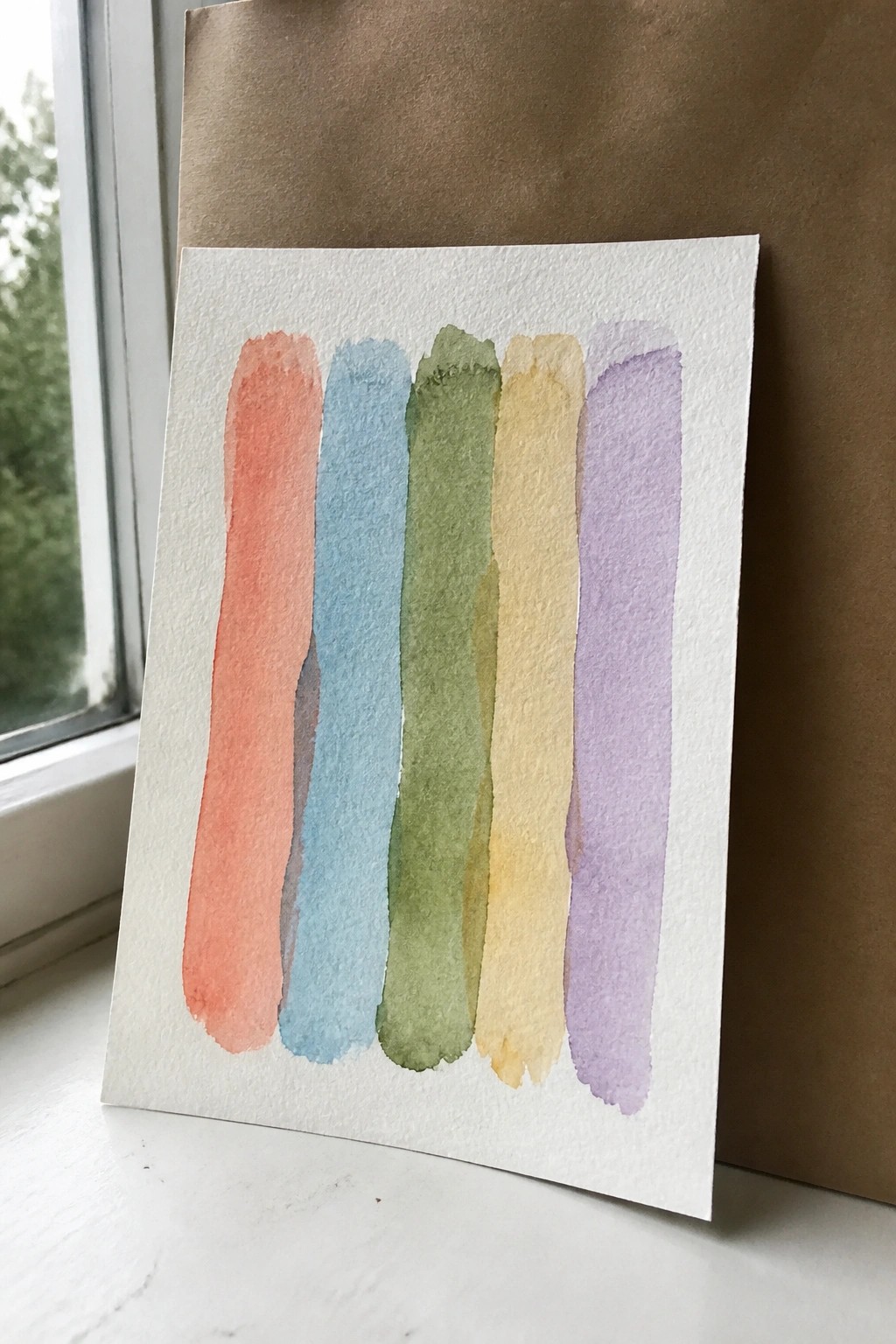 Five vertical watercolor stripes in coral, blue, green, yellow, and purple on white paper by window