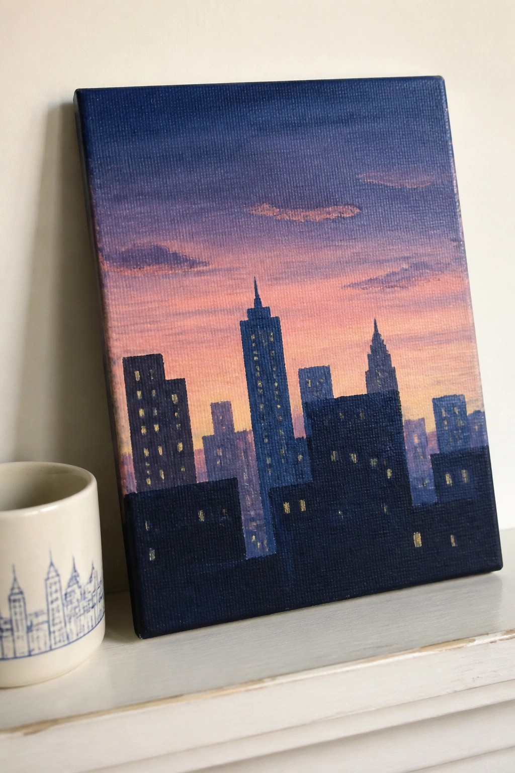 City skyline painting in blues and pinks with lit windows beside a white mug