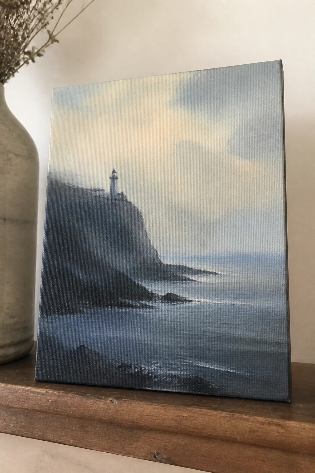 Painting of lighthouse on rocky cliff by misty sea beside ceramic vase on wood.