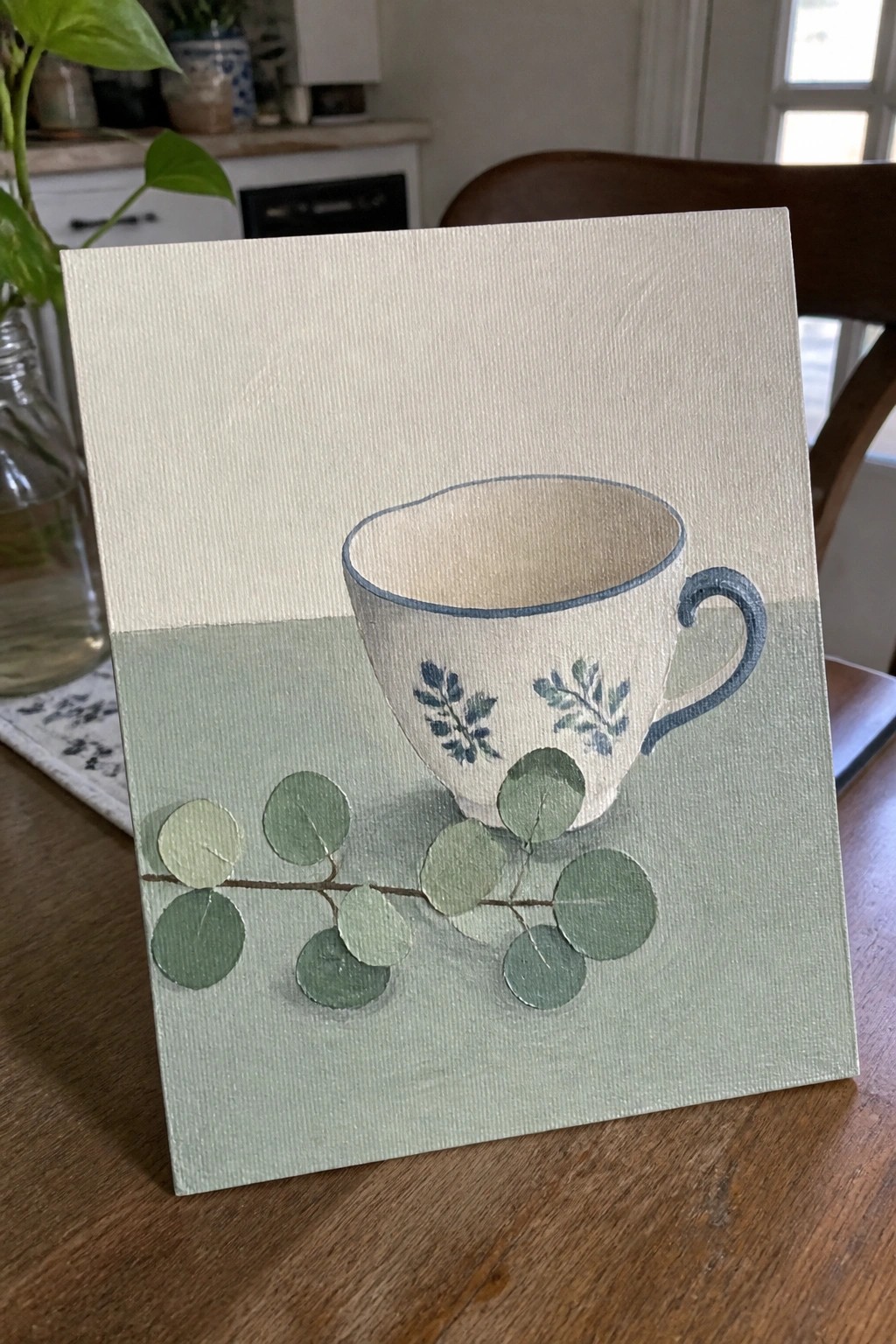 Small canvas painting of white teacup with blue trim and eucalyptus leaves
