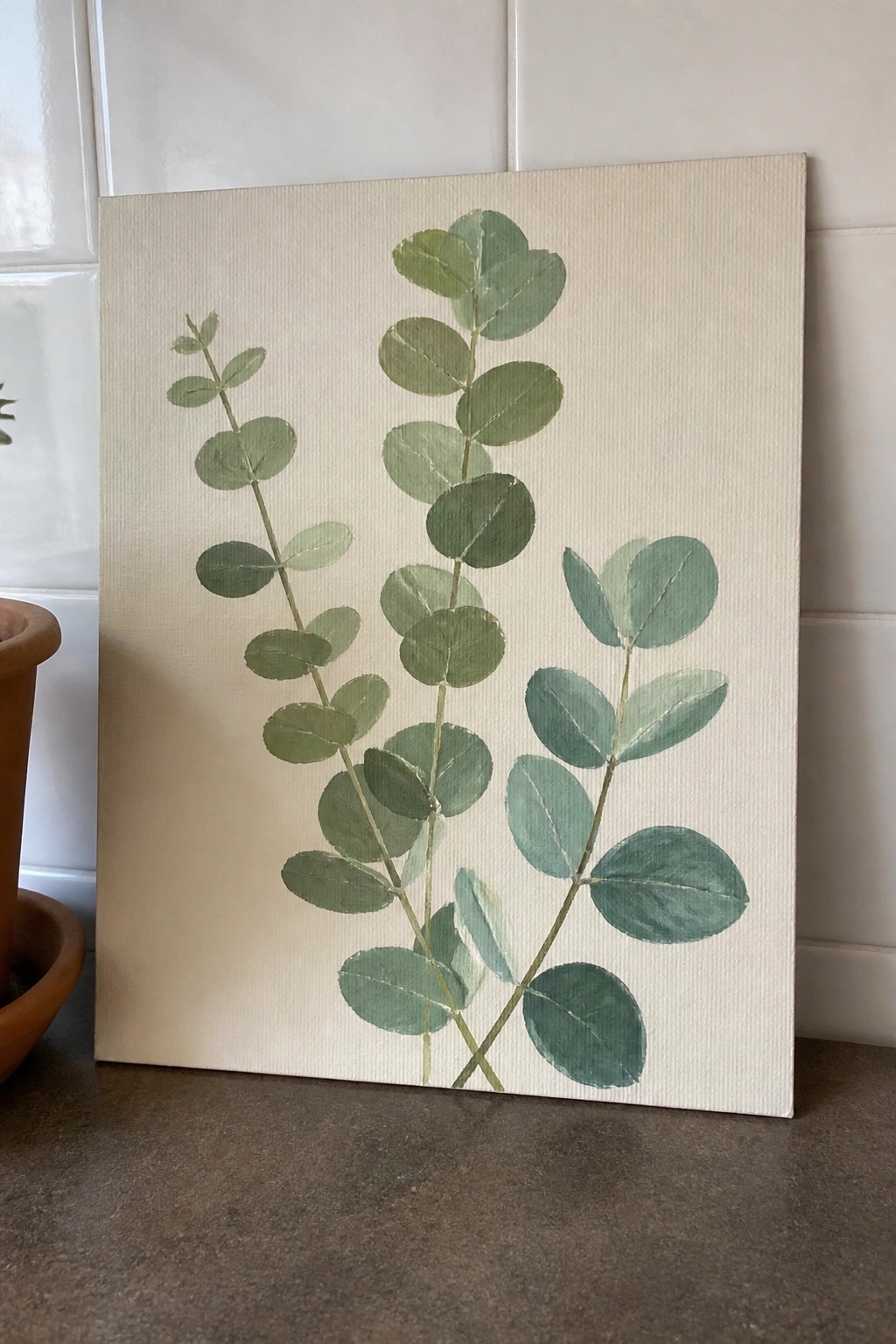 Green eucalyptus branches painted on canvas leaning against white tiled wall