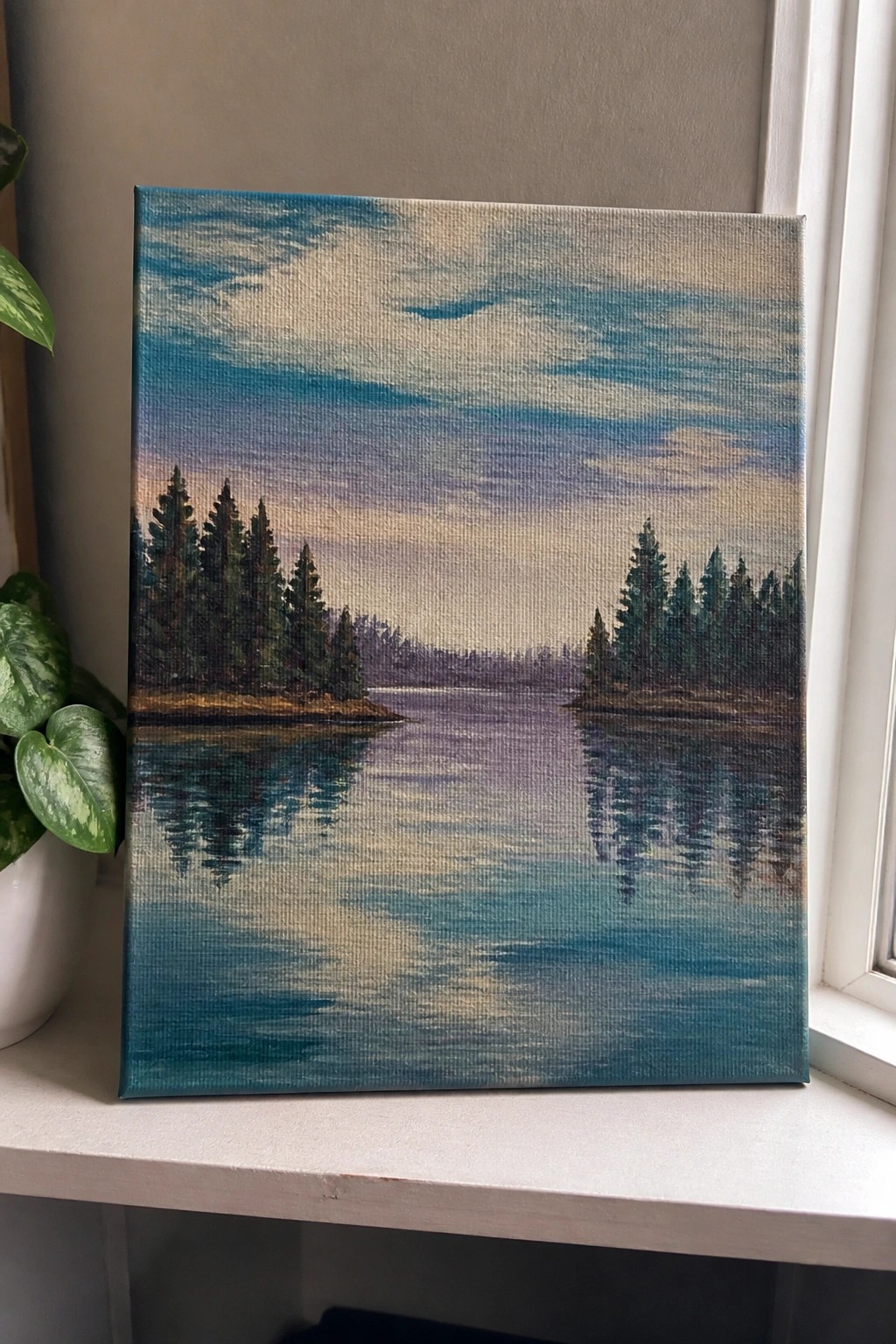 Painting of reflective lake with pine trees under colorful sky beside potted plant on windowsill.