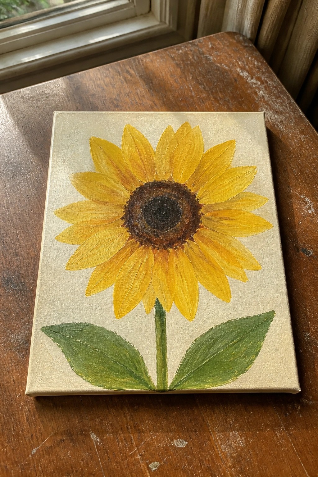 Yellow sunflower painting with brown center, green leaves on canvas atop wood table.