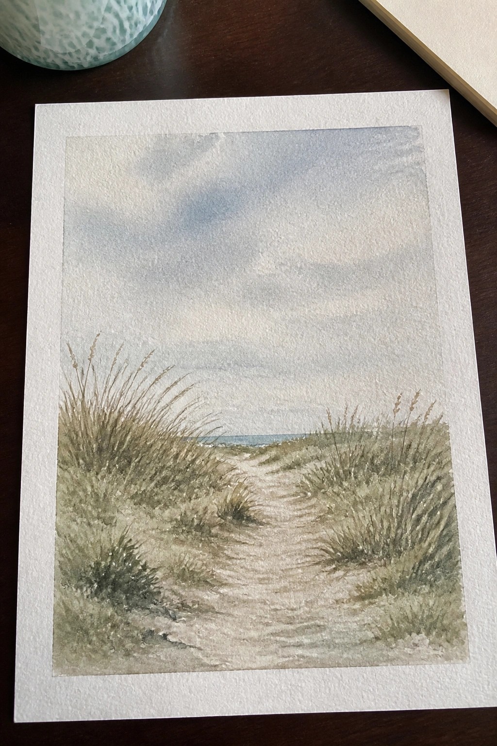 Watercolor painting of sandy path through grassy dunes leading to ocean under cloudy sky