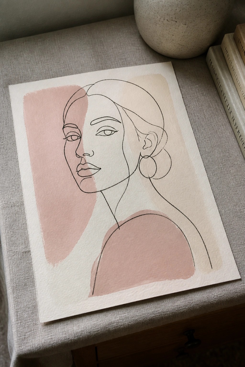 Minimalist line drawing of a woman on paper with pink shapes, beside vase and books