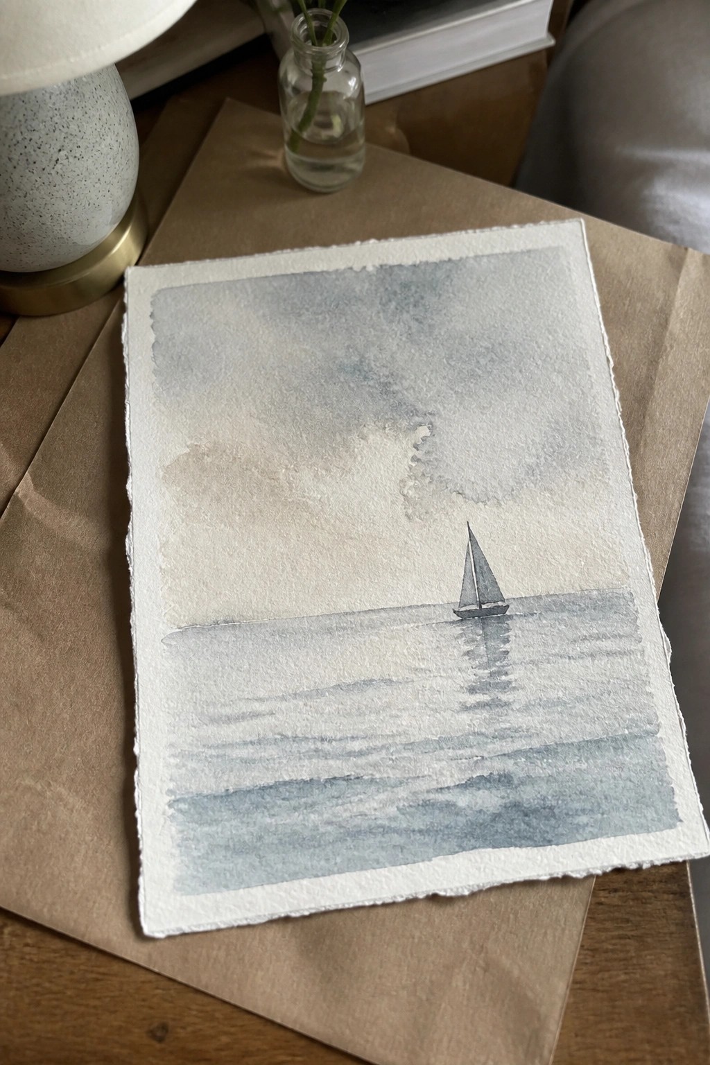 Watercolor of lone sailboat on calm blue waters beside speckled lamp and glass vase.
