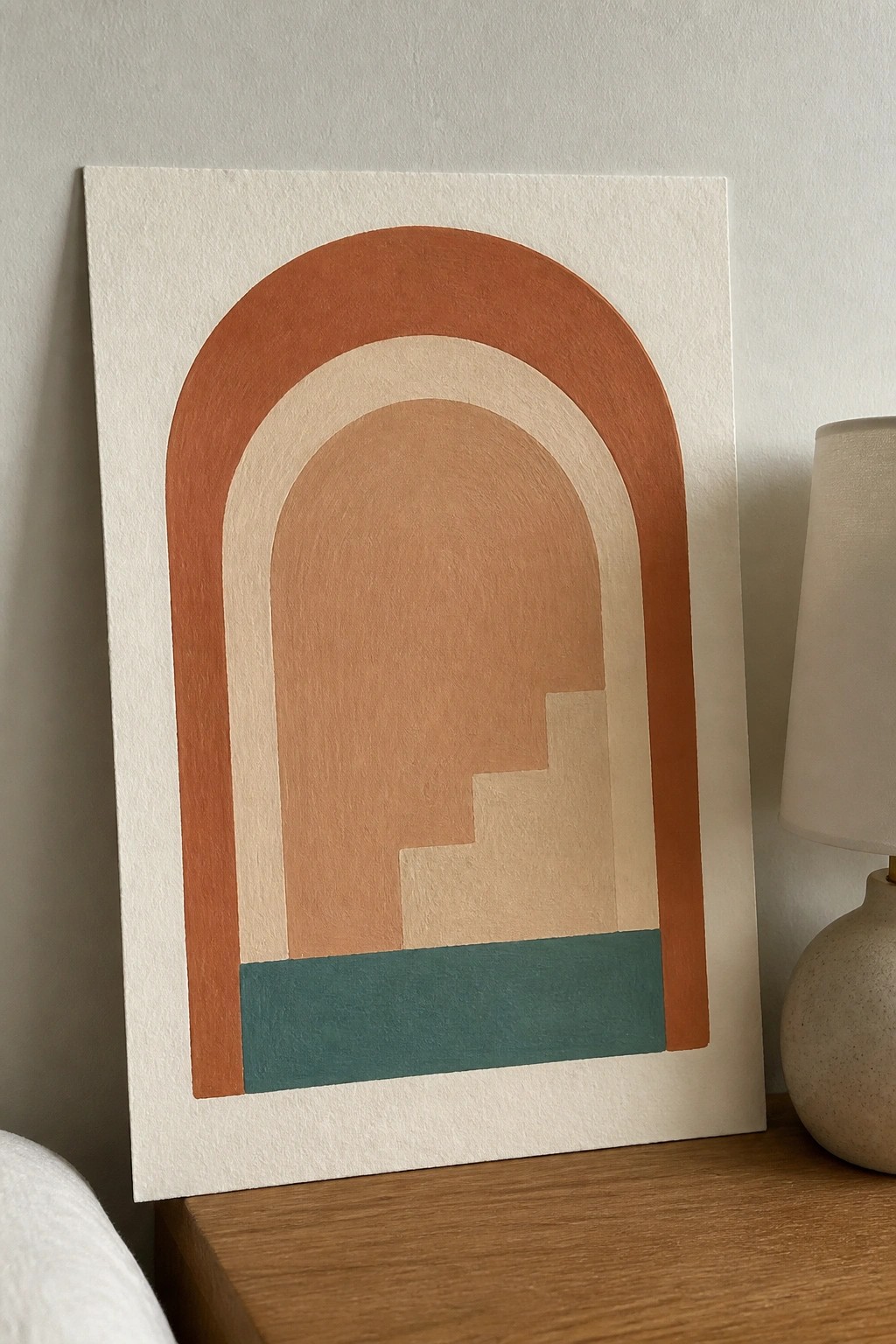 Abstract arched artwork in rust tones with teal base beside ceramic lamp on wood.