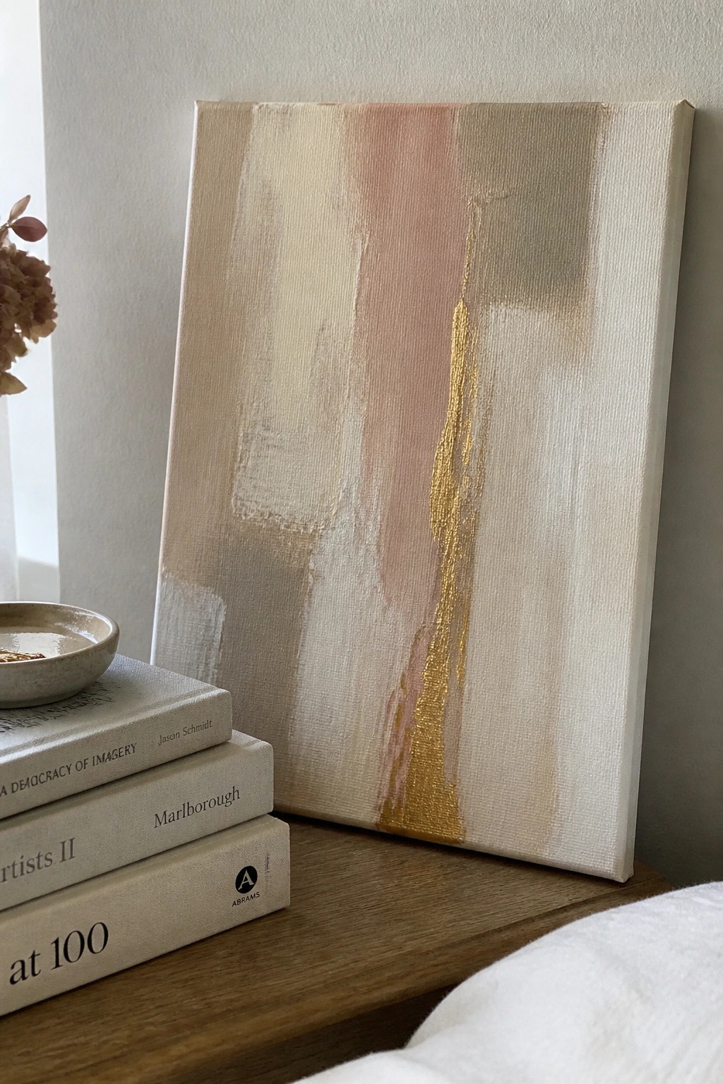 Abstract canvas with beige, pink, and gold strokes propped on books beside bowl.