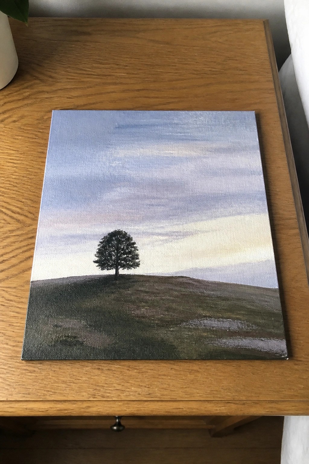 Lone tree painting on grassy hill under pastel sky, resting on wooden table.