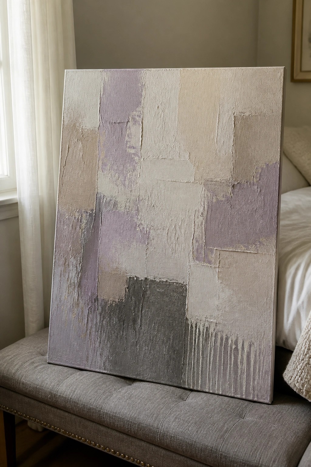 Abstract painting in soft neutrals and lavender leaning on gray upholstered bench.