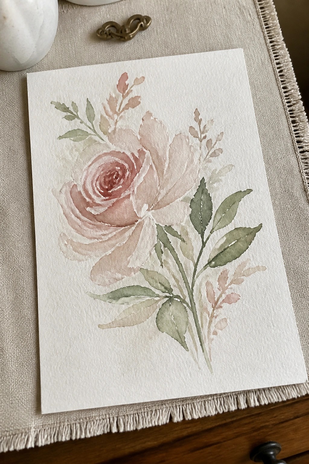 Watercolor painting of soft pink rose with green leaves on white paper over linen fabric