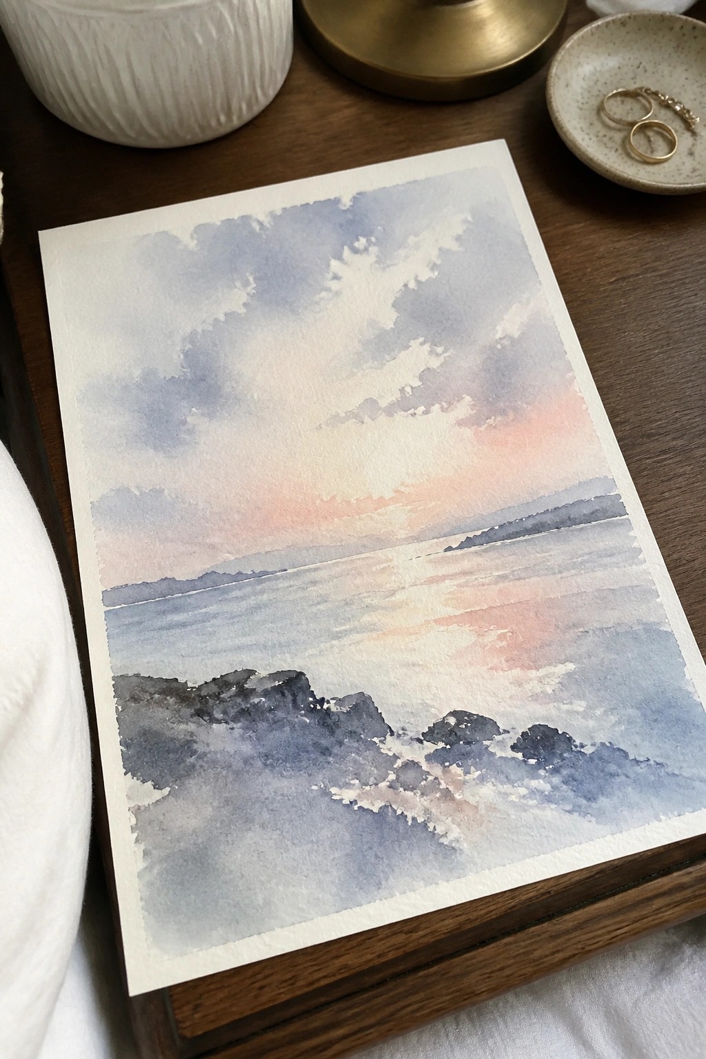Watercolor seascape of pink sunset over water with rocky shore on wooden table