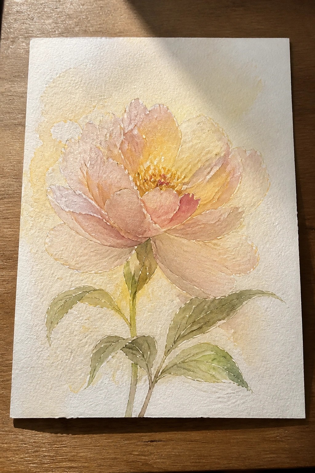 Watercolor peony in soft pink and yellow tones with green leaves and stem.
