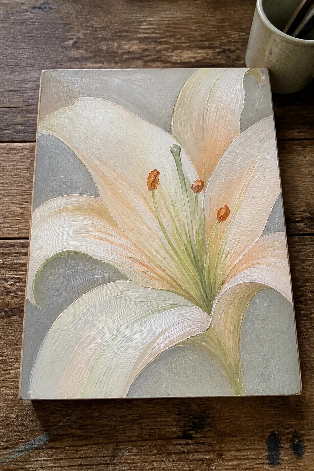 White lily painting with orange anthers on wooden table beside paintbrush cup