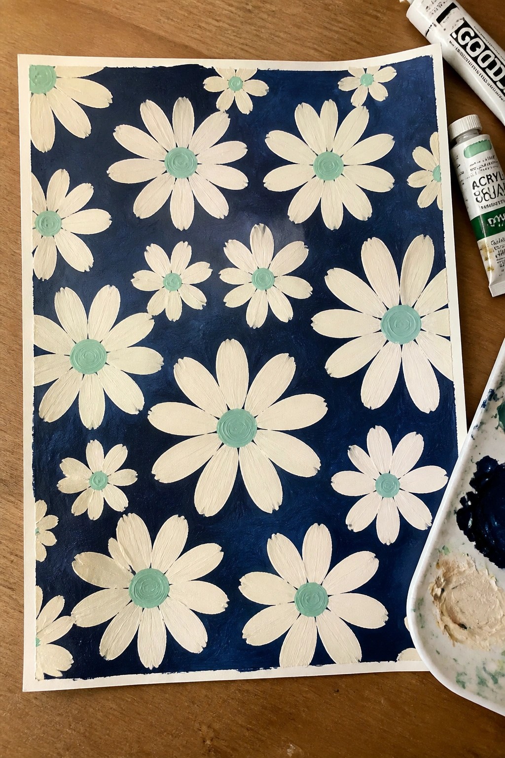 White daisies with green centers on dark blue background beside acrylic paint tubes and palette.