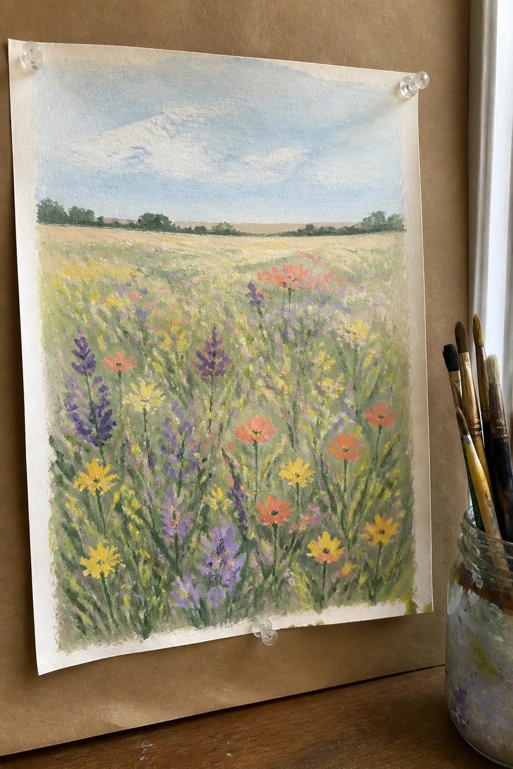 Watercolor wildflower meadow painting pinned to brown wall beside jar of brushes
