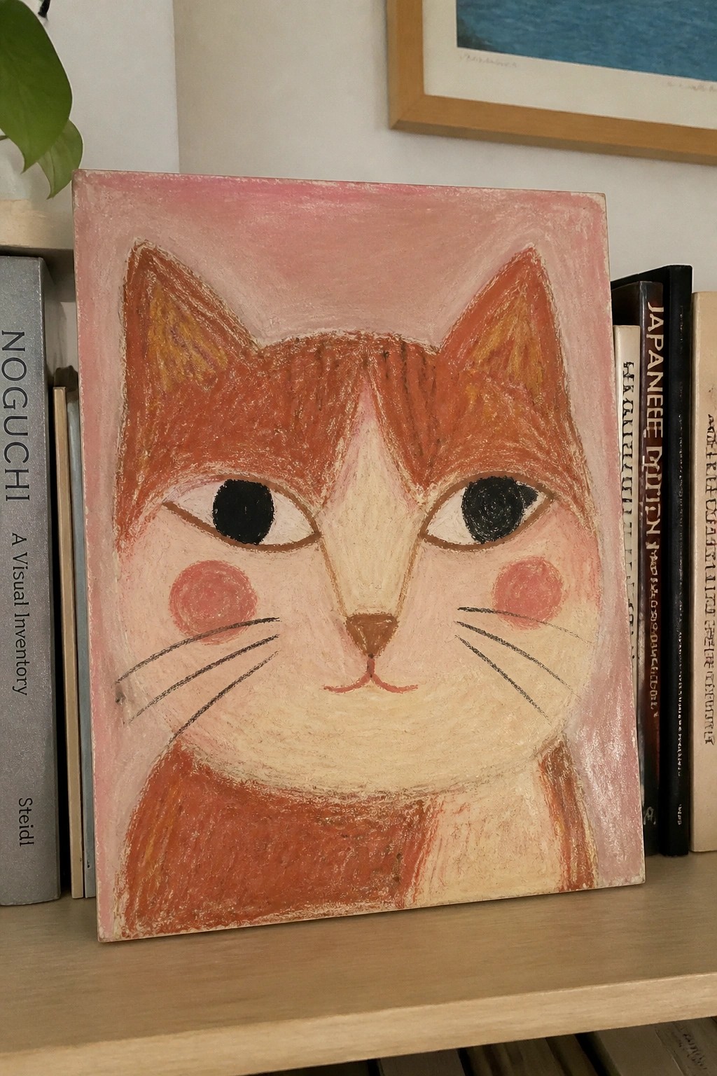 A painting of an orange cat face with large black eyes and pink cheeks against a pink background, leaning on a wooden shelf next to books.