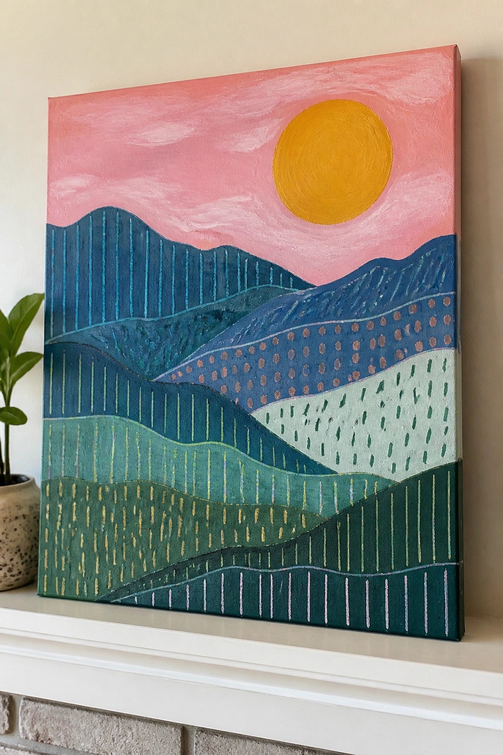 Abstract landscape painting of pink sky, yellow sun, and patterned hills on white mantel beside plant