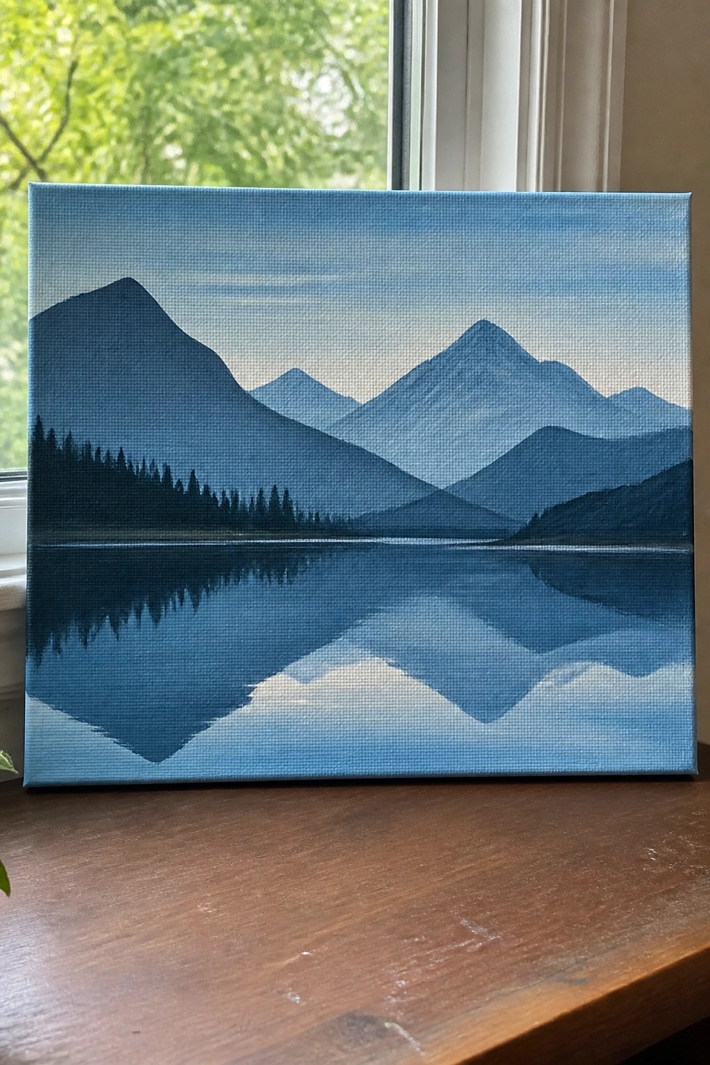 Layered blue mountains reflected in still lake, painted on canvas by window