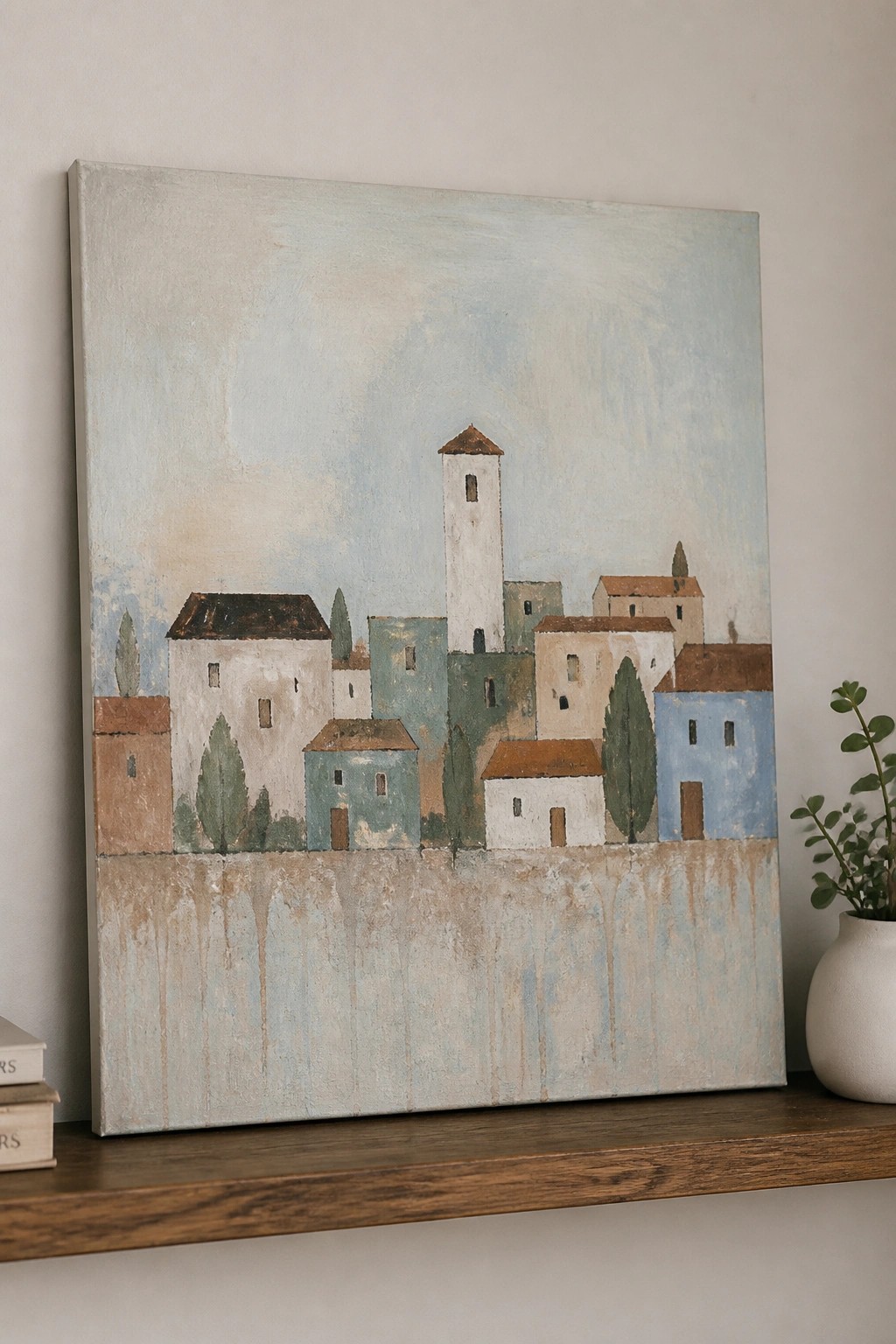 Pastel village painting with tower on wooden shelf beside books and vase.